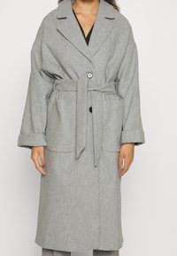 Vrouw in een lange grijze gebreide wollen jas met brede revers, twee grote voorzakken en knoopsluiting over een zwart topje.