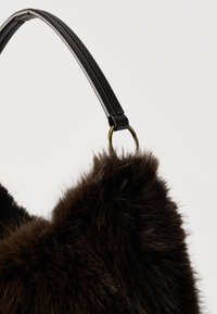 Σκούρο καφέ τσάντα με γούνινη υφή και μαύρο δερμάτινο λουρί που είναι προσαρτημένο με μπρούτζινο κρίκο, εμφανιζόμενο πάνω σε απλό ανοιχτό φόντο.