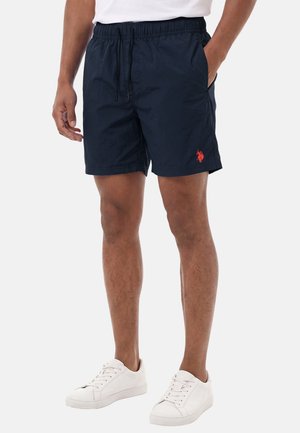 Mann trägt marineblaue Shorts mit rotem Logo, weiße Sneakers und ein weißes Hemd, steht mit einer Hand in der Tasche.