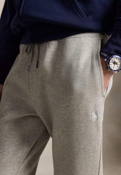 Personne portant un pantalon de survêtement gris clair avec une poche latérale et un sweat-shirt bleu marine, main dans la poche, montre visible au poignet.