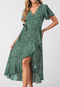 Vestido envolvente verde con lunares blancos, mangas cortas con volantes, dobladillo asimétrico y un lazo en la cintura para un ajuste ajustable. Fabricado en un tejido ligero.