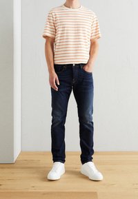 Camiseta a rayas en crema con rayas naranjas, combinada con jeans azul oscuro y zapatillas blancas. Ajuste casual, manga corta y diseño de cuello redondo.