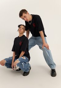 Zwei junge Erwachsene tragen schwarze T-Shirts mit Aufdrucken und zerrissene Jeans und posieren vor einem schlichten hellen Hintergrund, einer hockt und der andere lehnt sich nach vorne.