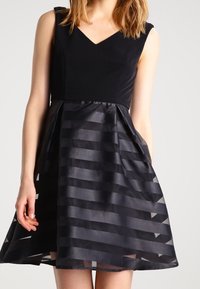 Svart klänning med en figursydd bodice och en utsvängd kjol. Har en V-ringning och lager av transparent tyg med horisontella ränder. Slät textur.