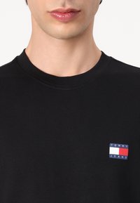 Crna pamučna majica s malim pravokobnim logom Tommy Jeans u plavoj, crvenoj i bijeloj boji na lijevoj strani prsiju. Okrugli izrez i kratki rukavi.