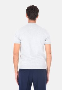 Uomo con i capelli corti e scuri, che indossa una camicia a maniche corte grigia chiara e pantaloni blu scuro, di spalle contro uno sfondo semplice.