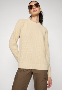 Beige gebreide trui met een gestructureerd patroon, geribde kraag en manchetten, gecombineerd met bruine cargobroek. Heldere achtergrond.