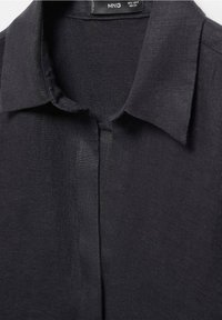 Chemise en lin noire avec un col classique, une fermeture par boutons sur le devant et une texture lisse. Coutures détaillées visibles au niveau du col et de la patte de boutonnage.