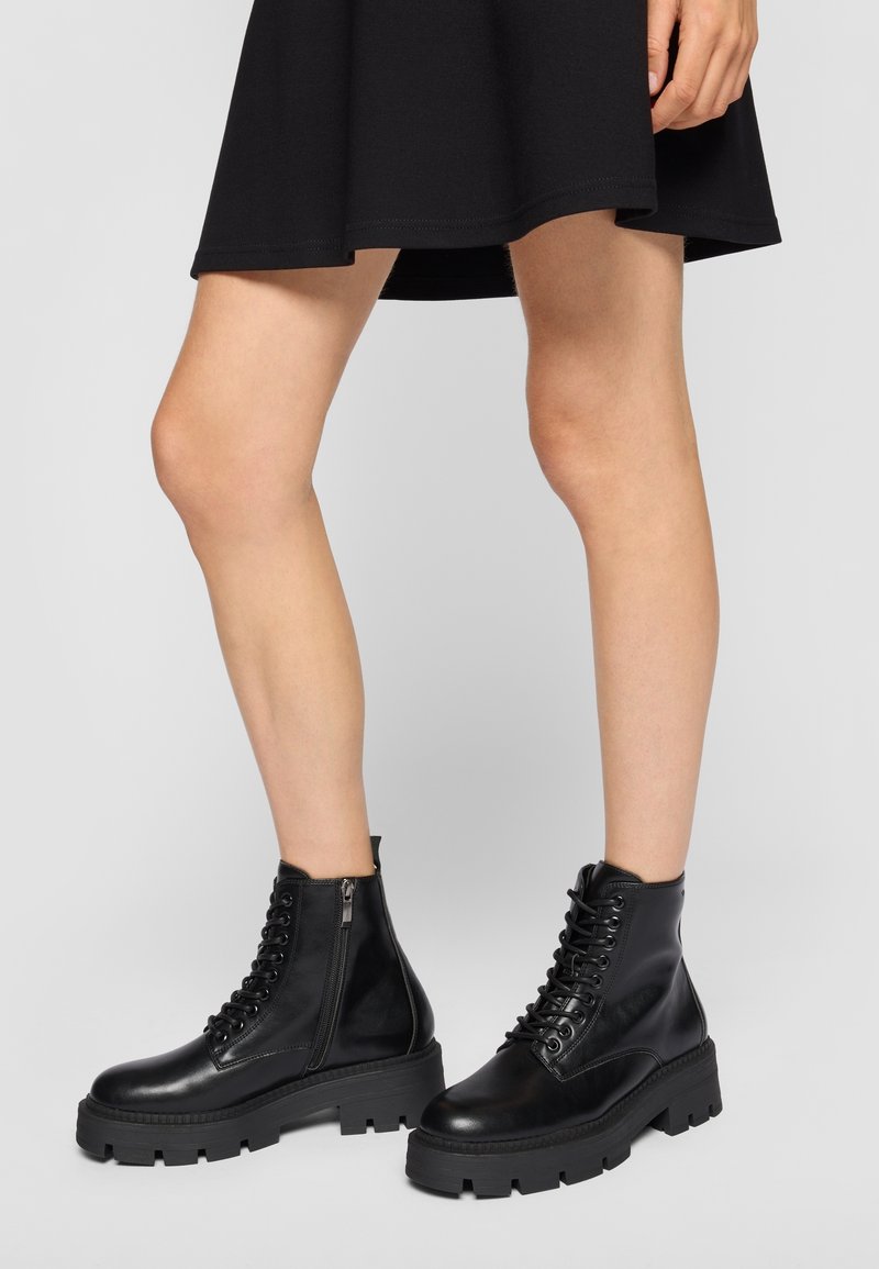 Jambes portant des bottines noires épaisses à lacets avec fermetures éclair latérales et une courte jupe noire sur un fond uni.