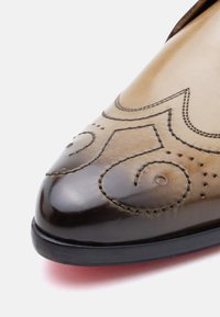 Καφέ δερμάτινο παπούτσι με μυτερή μύτη, με διακοσμητικά σπειροειδή σχέδια, ραφές ως διακοσμητικά στοιχεία και γυαλιστερό φινίρισμα. Μαύρη επίπεδη σόλα.