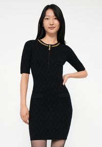 Jeune femme portant une robe en tricot texturé noire avec des manches courtes, des poches avant et un détail de fermeture éclair en chaîne dorée au niveau du col.