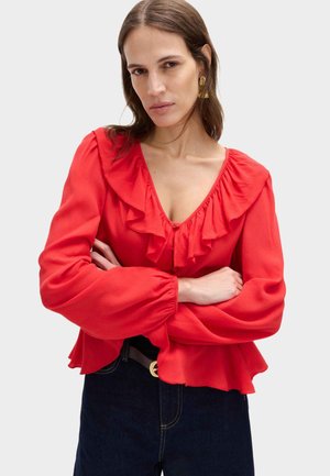 Kobieta z długimi brązowymi włosami, nosząca złote kolczyki i czerwoną bluzkę z falbanami, z założonymi rękami na jasnym, jednolitym tle.