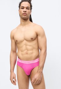 Μυώδης άνδρας με κράμπες φοράει φωτεινά ροζ εσώρουχα Calvin Klein, στέκεται μπροστά σε απλό ανοιχτό γκρι φόντο.