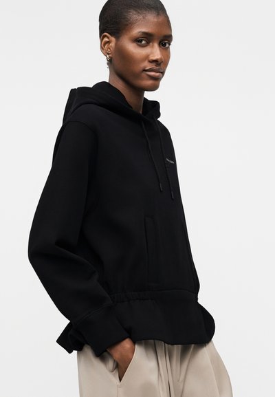 Γυναίκα με σκούρο δέρμα που φοράει μαύρο φούτερ με κουκούλα και μπεζ παντελόνι, στέκεται με τα χέρια στις τσέπες, πάνω σε απλό λευκό φόντο.