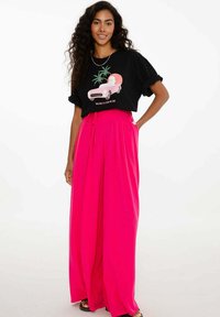 Žena s dlouhými kudrnatými vlasy má na sobě černé tričko s grafikou a jasně růžové široké kalhoty. Stojí s jednou rukou v kapse a usmívá se.