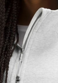 Sweat-shirt gris clair à fermeture éclair argentée et col montant, avec une texture douce et une coupe ajustée. Cheveux tressés visibles sur le côté.