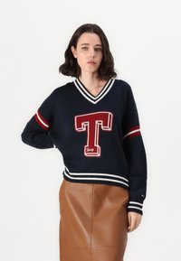 Maglione lavorato a maglia blu navy con logo rosso "T", strisce bianche sui polsini e sull'orlo, e scollo a V, abbinato a una gonna di pelle marrone.