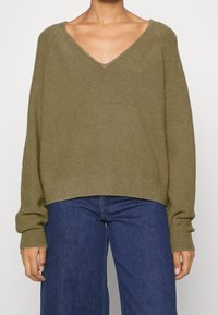 Olivgrüner, oversized Pullover mit tiefem V-Ausschnitt, gerippten Bündchen und geradem Saum. Weicher, strukturierter Stoff; kombiniert mit blauen weit geschnittenen Jeans.