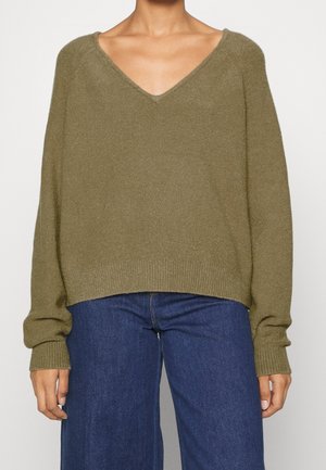 Pull oversized vert olive avec un décolleté en V profond, des poignets côtelés et un ourlet droit. Tissu doux et texturé ; porté avec un jean bleu à jambe large.