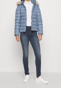 Chaqueta de plumas azul clara con capucha con borde de pelo, puños acanalados y cierre con cremallera; combinada con jeans ajustados oscuros y zapatillas blancas.
