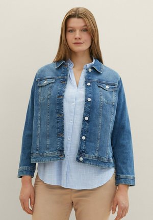 Giacca di jeans blu, con tasche frontali, chiusura a bottoni e un design leggermente corto. Indossata sopra una camicia azzurra chiara.