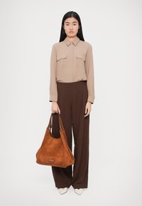 Béžová košile na knoflíky se dvěma kapsami na prsou, v kombinaci s hnědými kalhotami s širokými nohavicemi. Drží hnědou semišovou tašku na rameno.