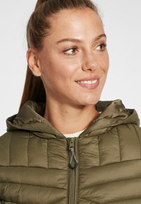 Olivenfarget polstret jakke med hette, kviltet design og frontglidelås med metalltrekk. Myk innerside synlig ved kragen.