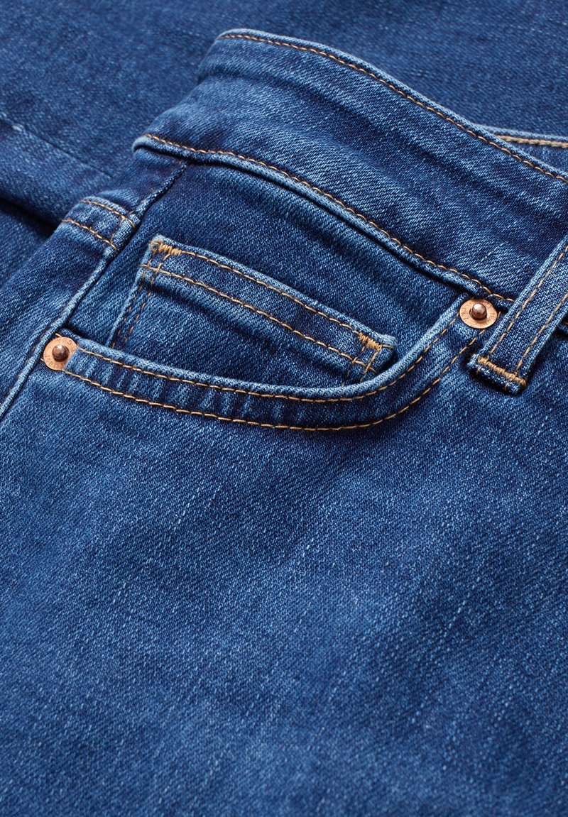 Blauer Denim-Stoff mit einer Nahaufnahme einer Vordertasche, die einen Kupferknopf und goldene Nähte zeigt, und eine glatte Textur sowie ein klassisches Design präsentiert.