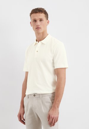 Cream-colored polo shirt made of textured fabric, with a buttoned placket and short sleeves, paired with light gray trousers.