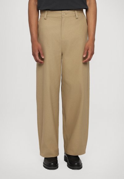 Beige, bő szárú nadrág sima felülettel, elülső gombos záródással és övhurkokkal, fekete lábbelivel kombinálva.