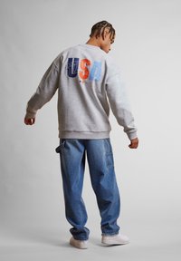 Felpa grigia con grafica multicolore "SISTER" sulla parte posteriore, abbinata a pantaloni cargo in denim blu a vestibilità morbida e sneakers bianche.