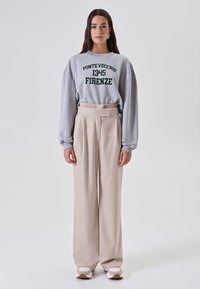 Donna in piedi che indossa un felpa grigia con la scritta "Ponte Vecchio 1345 Firenze", pantaloni beige a gamba larga e sneaker bianche su uno sfondo semplice.