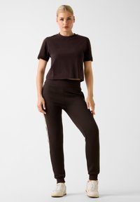 Kobieta z blond włosami w niskim kokie, ubrana w ciemno-brązowy krótki T-shirt i dopasowane dresy z bocznymi paskami, stojąca na tle w jednolitym kolorze.