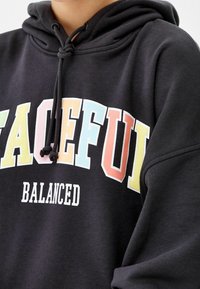 Czarna bluza z kapturem i sznurkiem. Napis „PEACEFUL” wielokolorowymi wielkimi literami oraz „BALANCED” w kolorze białym poniżej. Gładka faktura tkaniny.