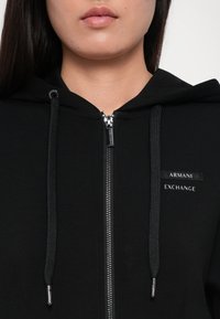 Gros plan d'une personne portant un sweat à capuche zippé noir Armani Exchange avec cordons et logo visible sur la poitrine.