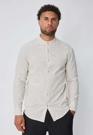 Homme aux cheveux bouclés et à la barbe, portant une chemise beige à manches longues avec col mandarin et un pantalon noir.