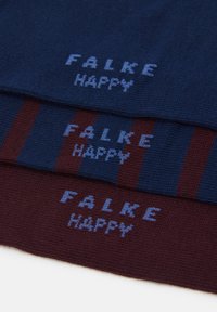 Tres calcetines muestran "FALKE HAPPY" en azul sobre un fondo de color marino y burdeos con un diseño de punto texturizado. El calcetín del medio presenta rayas.