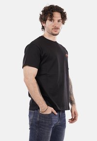 Maglietta nera in cotone a maniche corte, con scollatura a girocollo e un discreto dettaglio del logo arancione sul petto, abbinata a jeans blu.