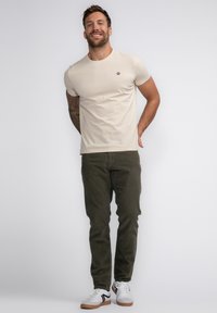 Beige t-shirt met korte mouwen en logo op de borst, gecombineerd met olijfgroene corduroy broek en witte sneakers met zwarte accenten.