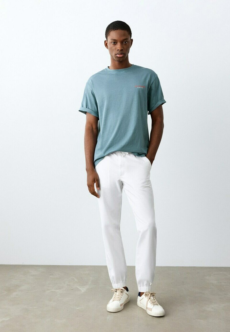 Jeune homme debout, portant un t-shirt bleu sarcelle à manches retroussées, un pantalon de jogging blanc et des baskets blanches, devant un mur uni clair.