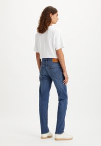 Oseba stoji obrnjen bočno, oblečena v belo majico s kratkimi rokavi, modre kavbojke z zadnjimi žepi in bele teniske z gumijastimi podplati.