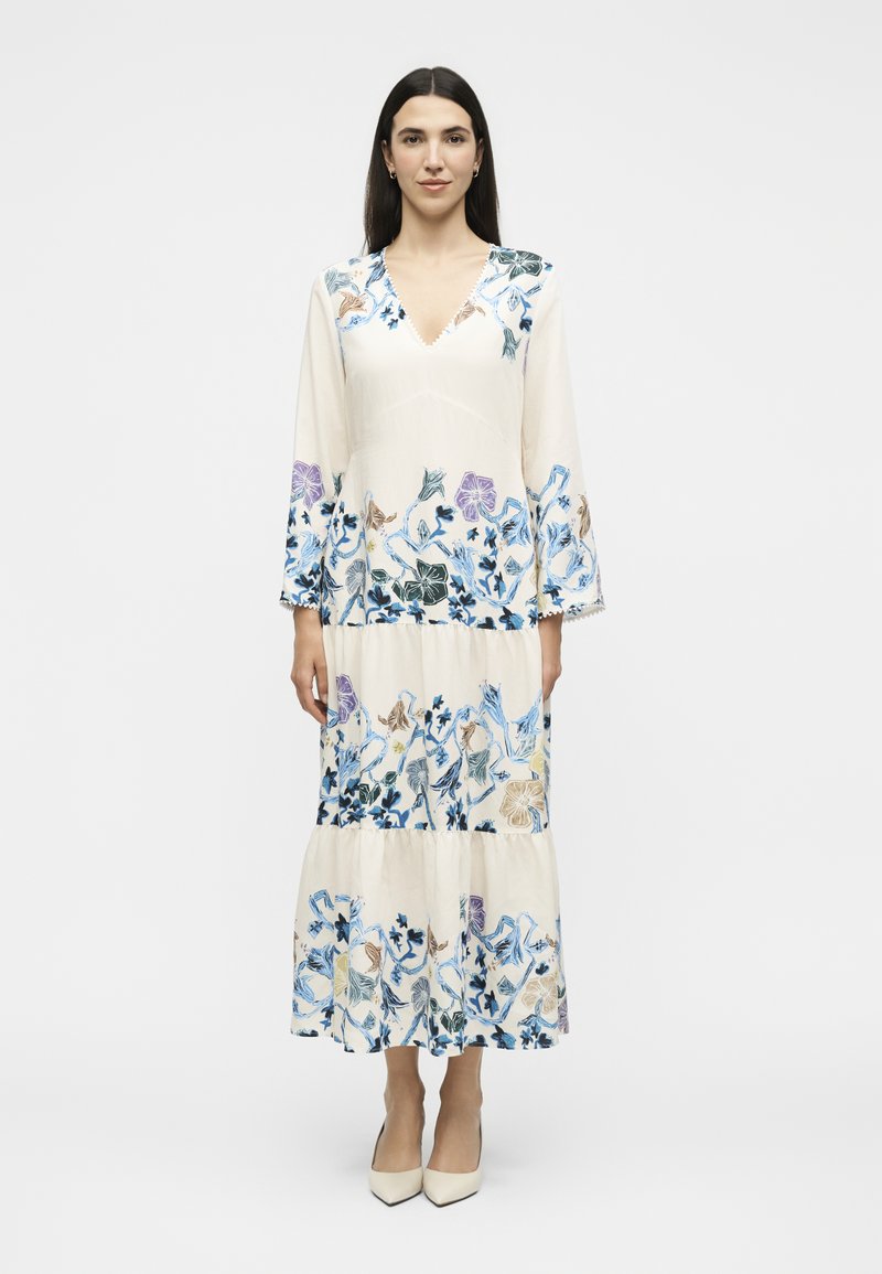 Femme debout, portant une longue robe blanche avec des motifs floraux bleus, violets et beiges, manches longues et talons blancs à bout pointu.