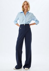 Camicia azzurra chiara con maniche arrotolate abbinata a pantaloni blu navy a vita alta con dettagli in bottone dorato e tasche frontali.