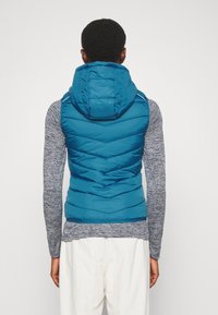 Türkiser Puffer-Weste mit Kapuze, ausgestattet mit einem Karo-Steppdesign und einem Reißverschluss vorne. Trägt man über einem langärmligen grauen Oberteil und hellen Hosen.