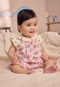 Salopetă roz pal cu imprimeu animal, guler cu dantelă și umeri cu volănașe; asortată cu șosete roz deschis, așezată pe un material bej.