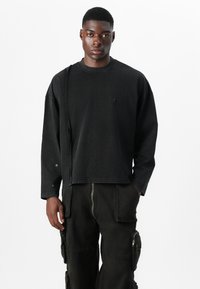 Zwart sweatshirt met lange mouwen en ronde hals, met een subtiel logo op de voorkant en een unieke stikselafwerking op één schouder.