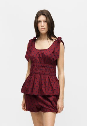 Jeune femme portant un haut sans manches bordeaux à pois bleu marine et un short assorti, debout devant un fond clair uni.