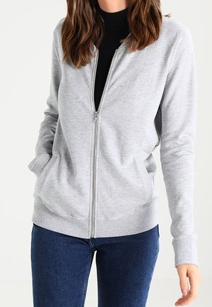 Woman wearing a light gray zip-up sweatshirt over a black top and blue jeans, with her left hand in the pocket and right arm relaxed.