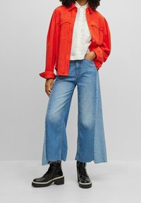 Röd denimskjorta, vit texturerad blus, vida blå jeans med split-design, och svarta snörstövlar med kraftiga sulor.