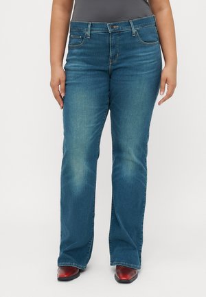 Personne portant un jean droit bleu et des bottes rouges à bouts pointus, debout devant un fond blanc.
